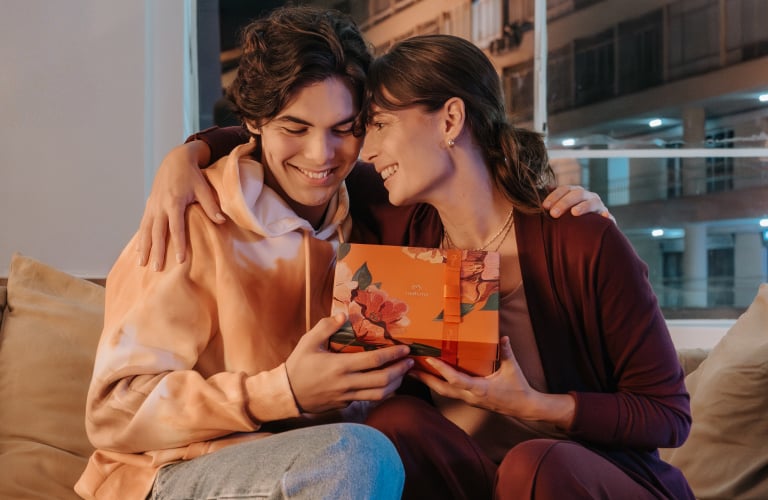 Foto em ambiente interno de uma mulher e um jovem sorrindo e abraçados no sofá, enquanto seguram juntos uma caixa de presente Natura.